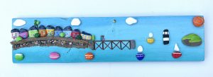 painted pebblescape depicting beaumaris' colourful houses, the pier with a couple on it flying a kite, boats sailing, penmon lighthouse and puffin island with the sun and billowing clouds in the sky by contemporary anglesey artist deborah kempton