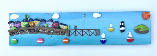 painted pebblescape depicting beaumaris' colourful houses, the pier with a couple on it flying a kite, boats sailing, penmon lighthouse and puffin island with the sun and billowing clouds in the sky by contemporary anglesey artist deborah kempton