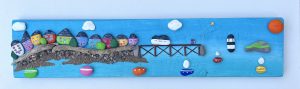 painted pebblescape depicting beaumaris' colourful houses, the pier with a couple on it flying a kite, boats sailing, penmon lighthouse and puffin island with the sun and billowing clouds in the sky by contemporary anglesey artist deborah kempton