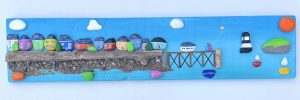 painted pebblescape depicting beaumaris' colourful houses, the pier with a couple on it flying a kite, boats sailing, penmon lighthouse and puffin island with the sun and billowing clouds in the sky by contemporary anglesey artist deborah kempton
